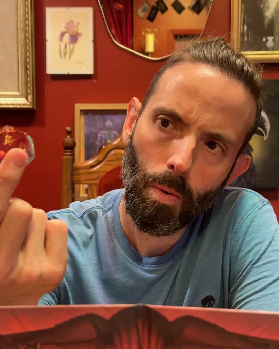 Daniel Rocha behind his hand-painted Game Master screen, holding up a red twenty-sided die, in a room hung with framed fantasy art
