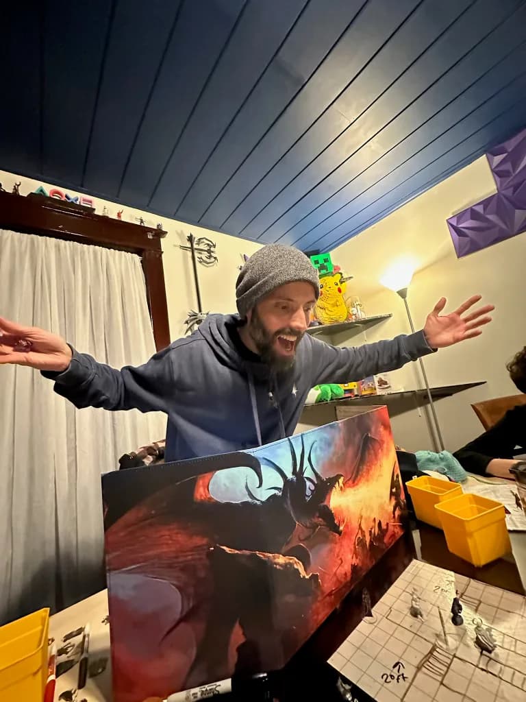Daniel Rocha behind his hand-painted dragon screen, arms thrown wide mid-scene, at a table laid with dice and miniatures